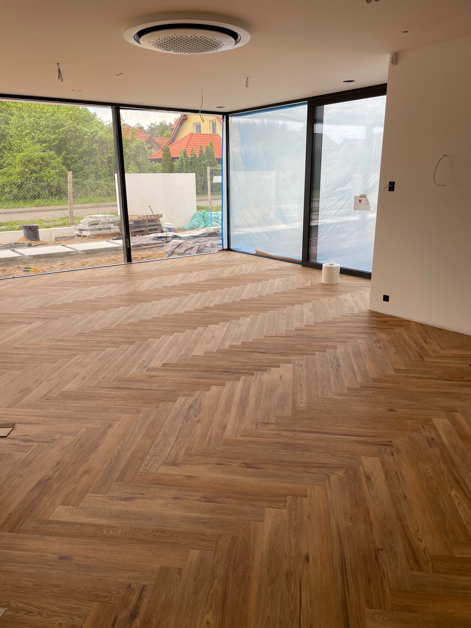 Nowoczesne wnętrze w trakcie wykańczania z parkietem ułożonym w jodełkę francuską, widoczne okna z czarnymi ramami i elementy instalacji sufitowej.