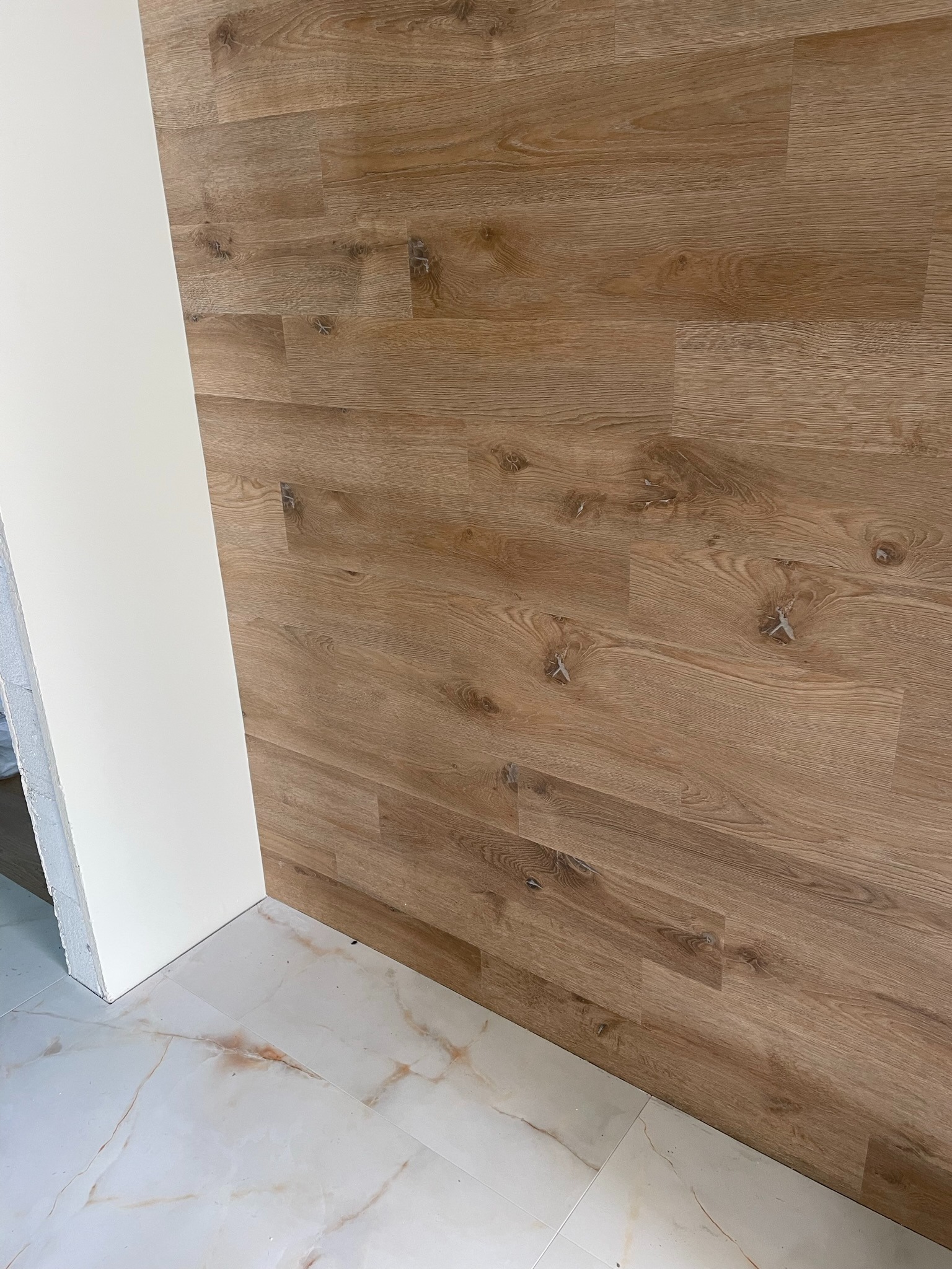 Panel ścienny imitujący drewno, ułożony poziomo, sąsiadujący z białą ścianą i marmurową podłogą.