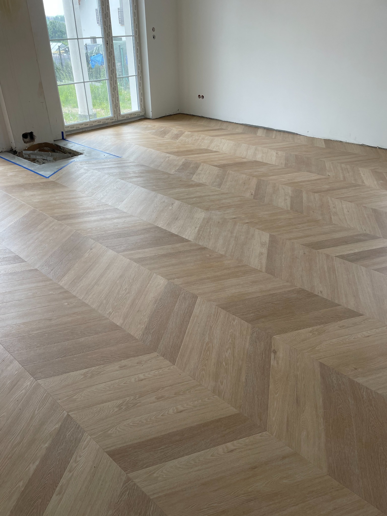Ułożona podłoga z jasnego drewna w jodełkę w nowym pomieszczeniu, z widocznym otworem w podłodze zabezpieczonym niebieską taśmą malarską przy ścianie z oknem.