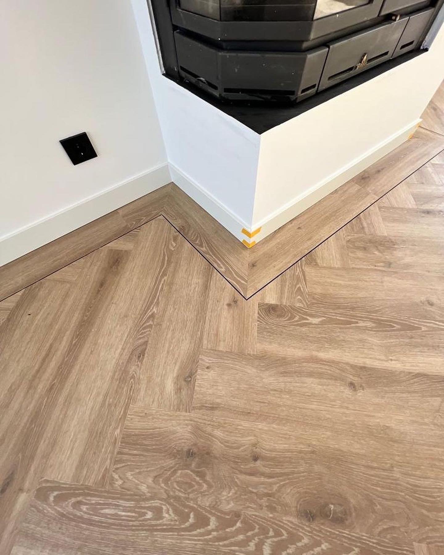 Ułożona parkietowa podłoga w jodełkę, jasne drewno, obudowa kominka z białymi listwami przypodłogowymi, czarny włącznik na ścianie.