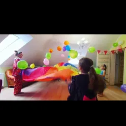 Kobieta w kostiumie klauna trzyma kolorową płachtę, wokół unoszą się balony, w tle dzieci i dekoracje urodzinowe w pokoju z oknem dachowym.