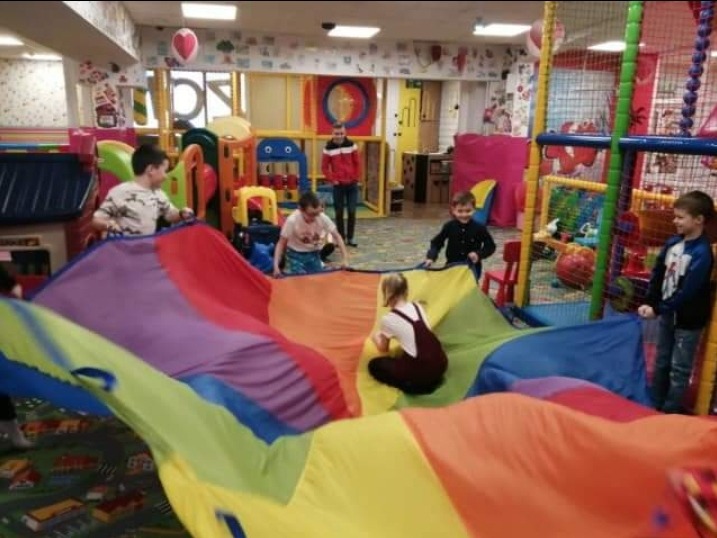 Grupa dzieci bawi się kolorową chustą animacyjną w sali zabaw z konstrukcjami do wspinaczki i dekoracjami ściennymi.