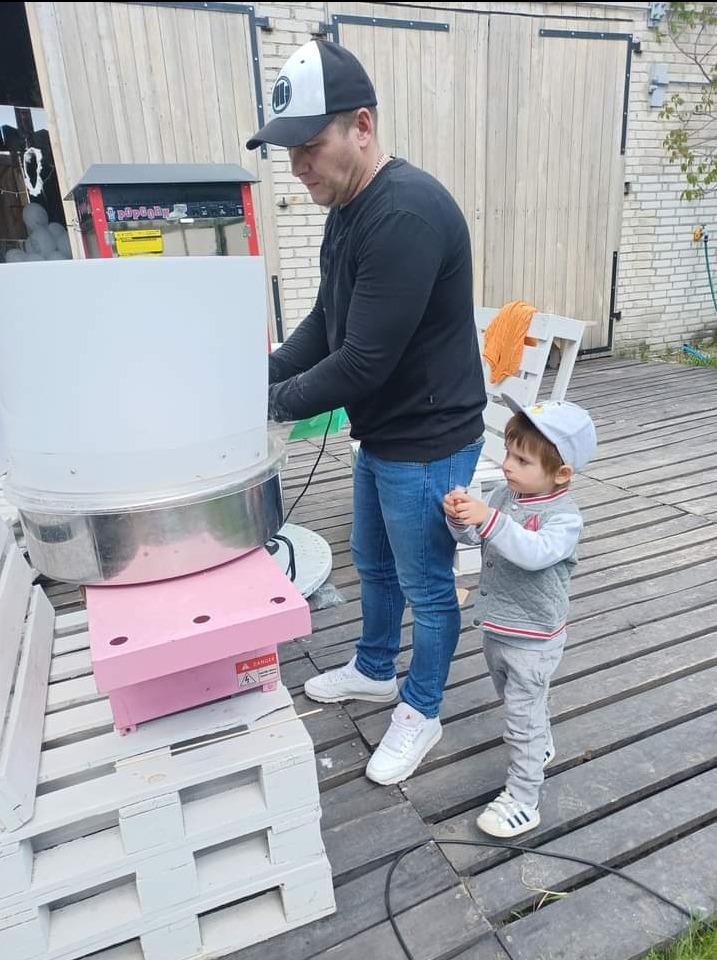 Mężczyzna w czapce obsługuje maszynę do waty cukrowej na drewnianym podeście, obok stoi mały chłopiec z watą w dłoniach.