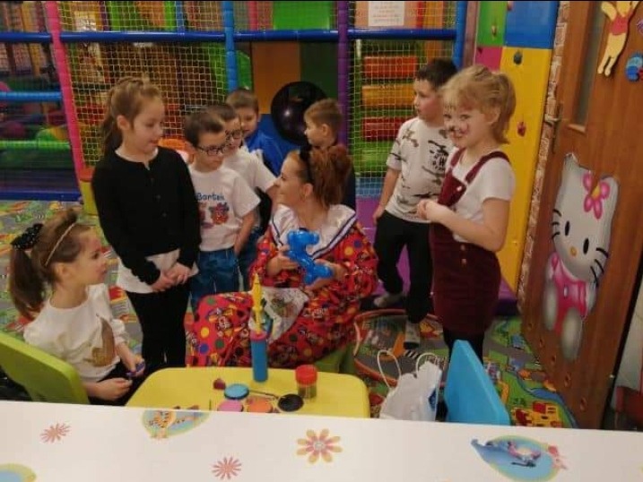 Kobieta w stroju klauna siedzi przy żółtym stoliku, trzymając niebieskiego balonowego dinozaura, otoczona grupą dzieci w sali zabaw.