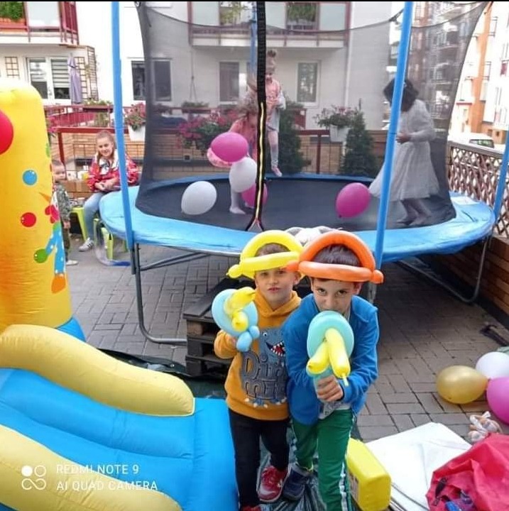 Dwaj chłopcy w balonowych kapeluszach trzymają balonowe pistolety na tle dmuchanego zamku i trampoliny z bawiącymi się dziećmi.