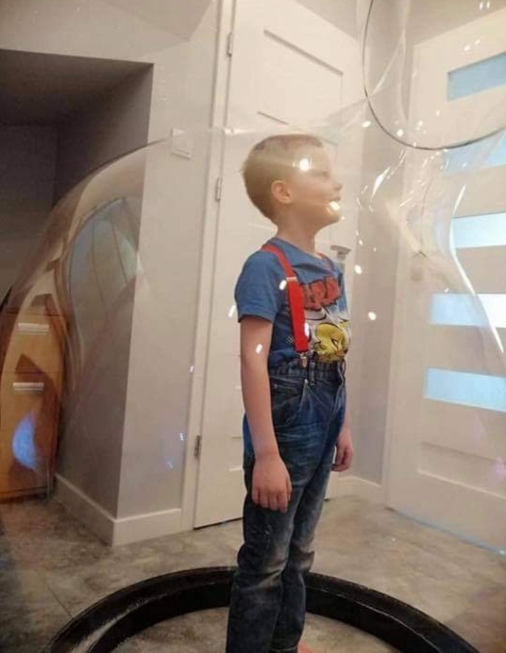 Chłopiec w dżinsowych ogrodniczkach z czerwonymi szelkami stoi wewnątrz dużej, przezroczystej bańki, z widoczną czarną podstawą i fragmentem drzwi w tle.