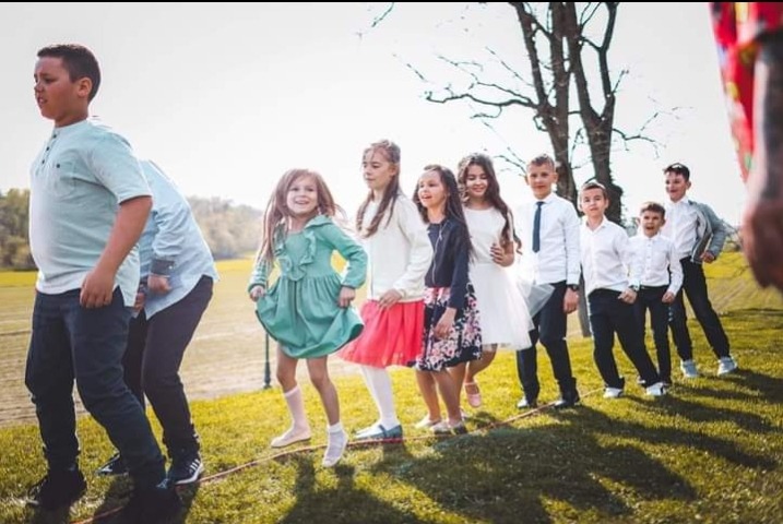 Grupa dzieci, chłopców i dziewcząt, ustawiona w rzędzie skacze przez skakankę rozciągniętą na trawie podczas zabawy na świeżym powietrzu.