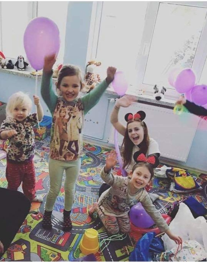 Animator z dziećmi na kolorowym dywanie z motywem ulic, dziewczynka w opasce z uszami Myszki Miki trzyma balon, w tle okno.