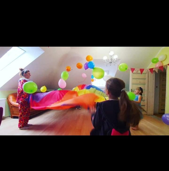 Kobieta w kostiumie klauna trzyma kolorową płachtę, wokół unoszą się balony, w tle dzieci i dekoracje urodzinowe w pokoju z oknem dachowym.