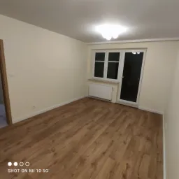 Pusty pokój po remoncie z panelami podłogowymi w odcieniach brązu, świeżo pomalowanymi ścianami w kolorze kremowym, białymi listwami przypodłogowymi, grzejnikiem pod oknem balkonowym i widocznym...