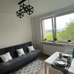 Salon z rozkładaną sofą, geometryczną lampą wiszącą i nakrytym stołem w jasnym wnętrzu z widokiem na miasto.