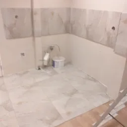 Niedokończona łazienka z częściowo położonymi płytkami imitującymi marmur na ścianach i podłodze; widoczne puszki instalacyjne, wiadro z farbą i fragment drabiny.