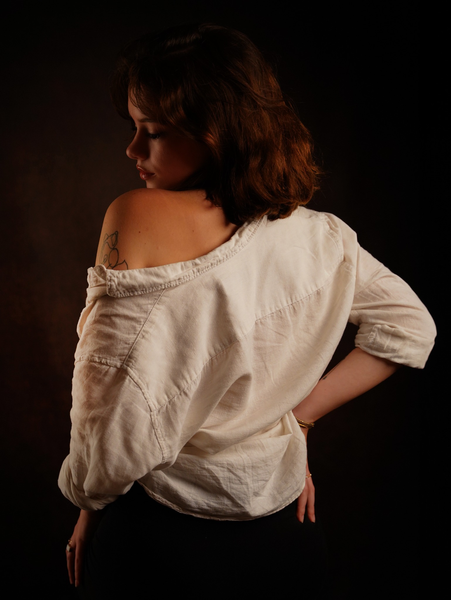 Portret kobiety odwróconej tyłem, w rozpiętej białej koszuli, z tatuażem na ramieniu i bransoletkami na nadgarstku, na ciemnym tle.