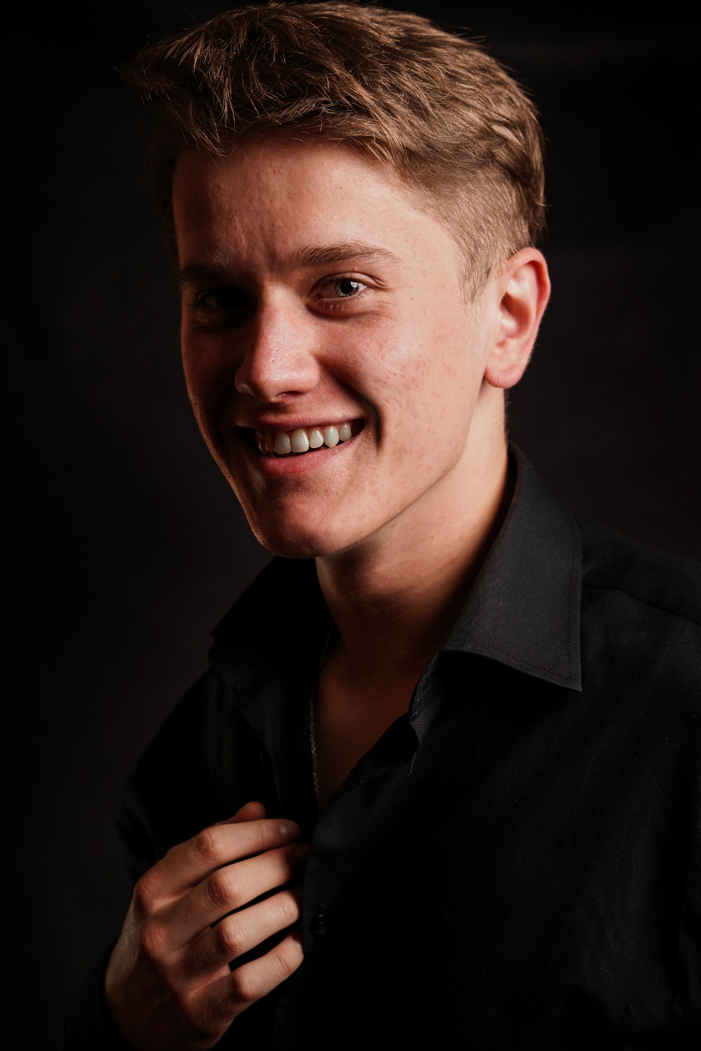 Młody mężczyzna w czarnej koszuli, uśmiechnięty, z ręką przy kołnierzyku, portretowe ujęcie na ciemnym tle.