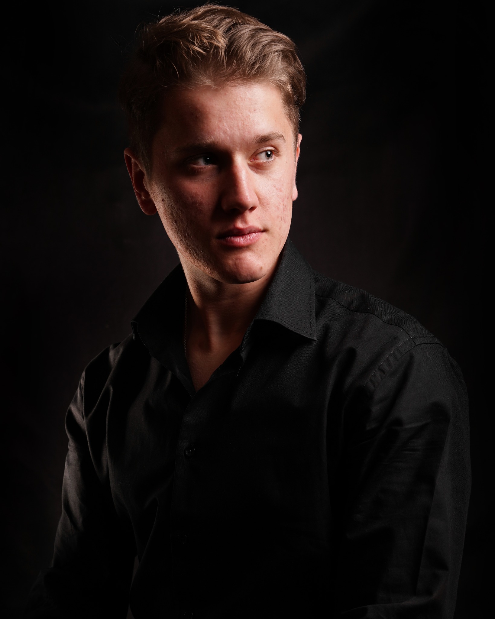Portret młodego mężczyzny w czarnej koszuli na ciemnym tle, z lekkim spojrzeniem w bok i widocznymi niedoskonałościami skóry.
