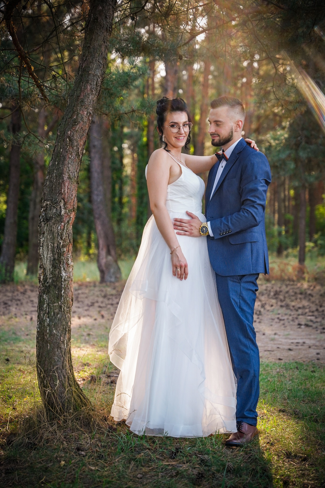 Para młoda w leśnej scenerii: Panna młoda w białej sukni z tiulowym dołem i okularach, pan młody w niebieskim garniturze i muszce, pozują przy drzewie w promieniach słońca.