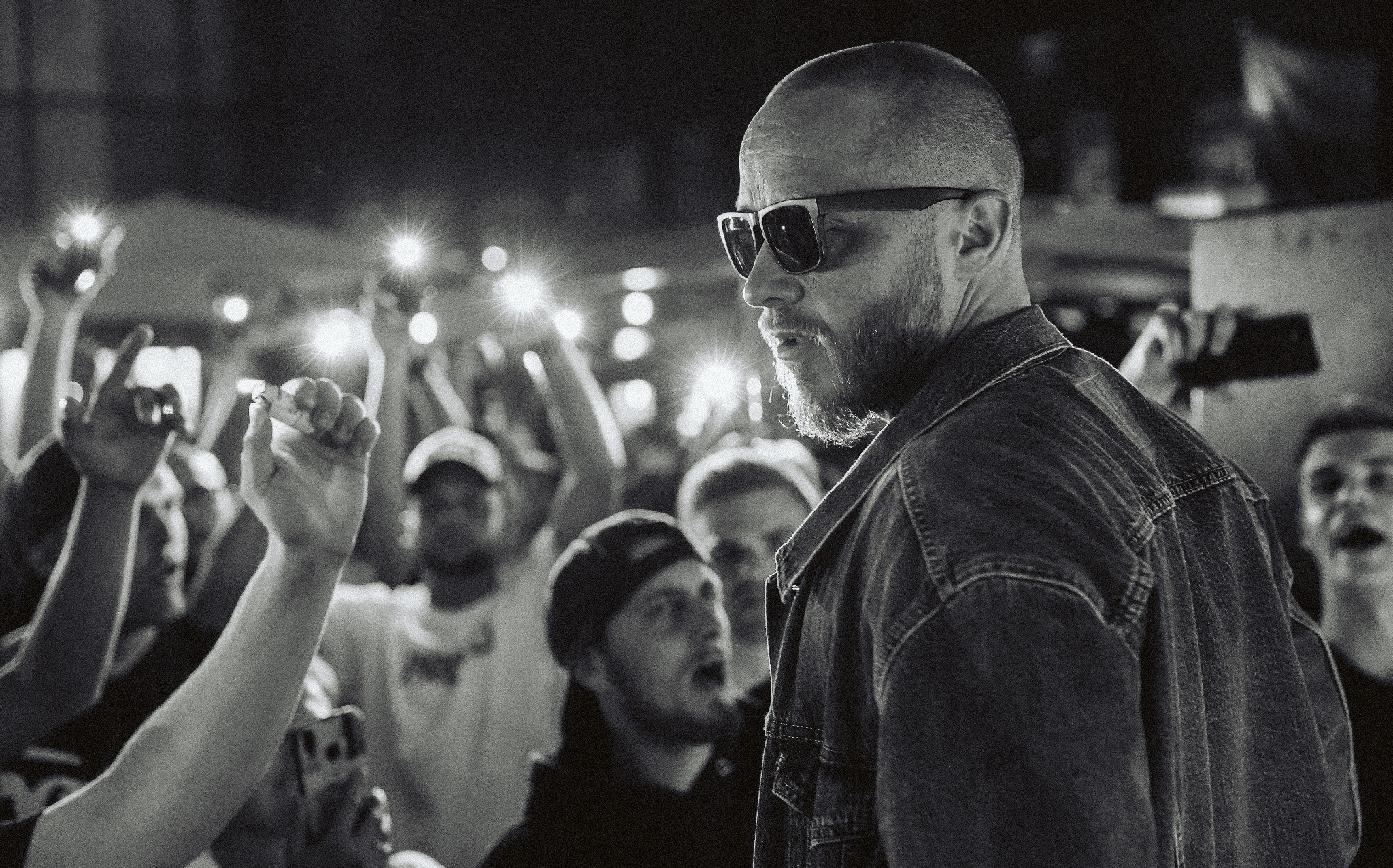 Czarno-białe zdjęcie z profilu mężczyzny w okularach na tle rozświetlonego tłumu z podniesionymi rękami, prawdopodobnie koncert.