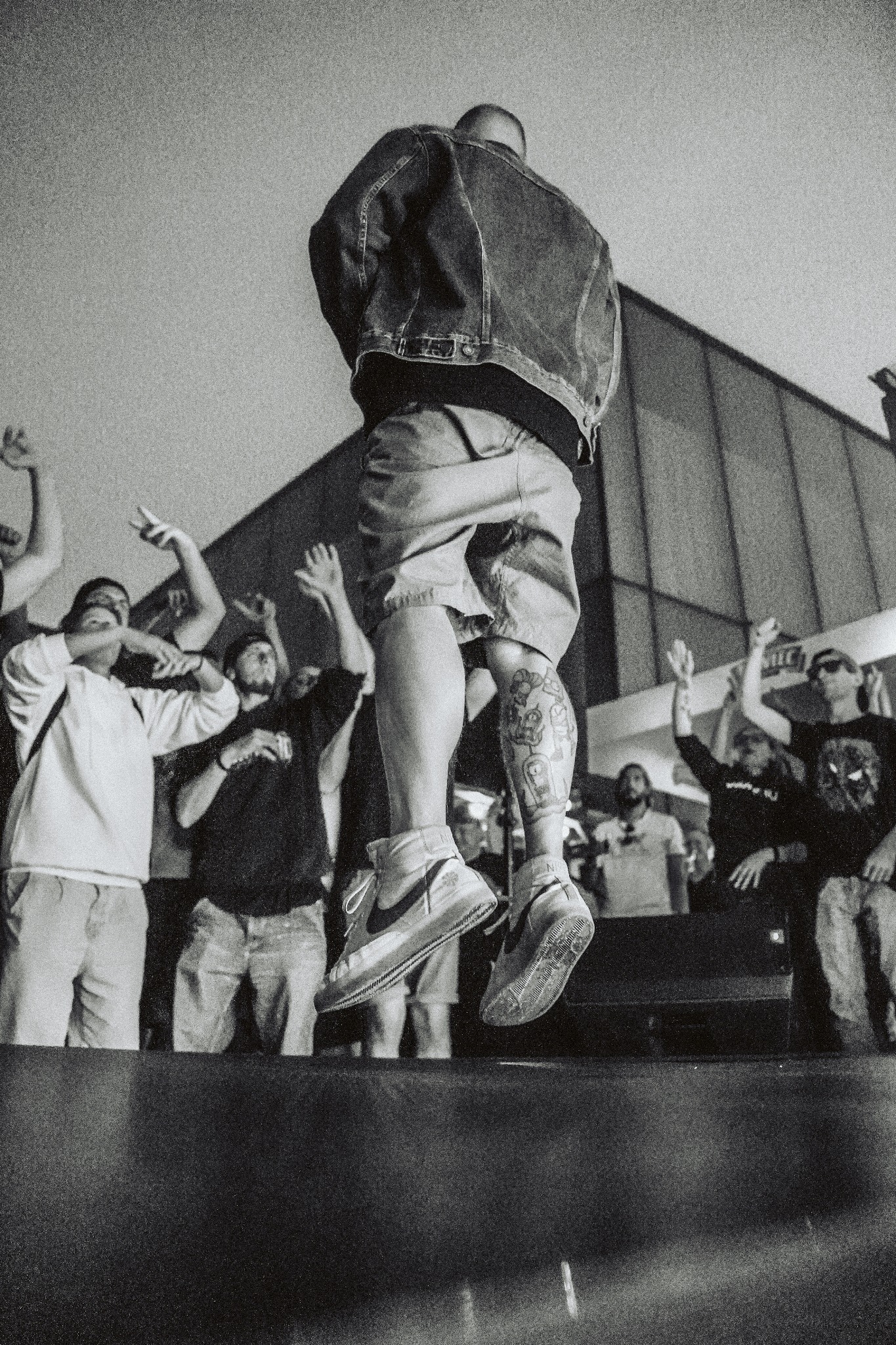 Czarno-białe zdjęcie skaczącego mężczyzny tyłem do aparatu, w dżinsowej kurtce i krótkich spodenkach, z tatuażem na łydce, podczas występu na scenie, z publicznością w tle z podniesionymi rękami.