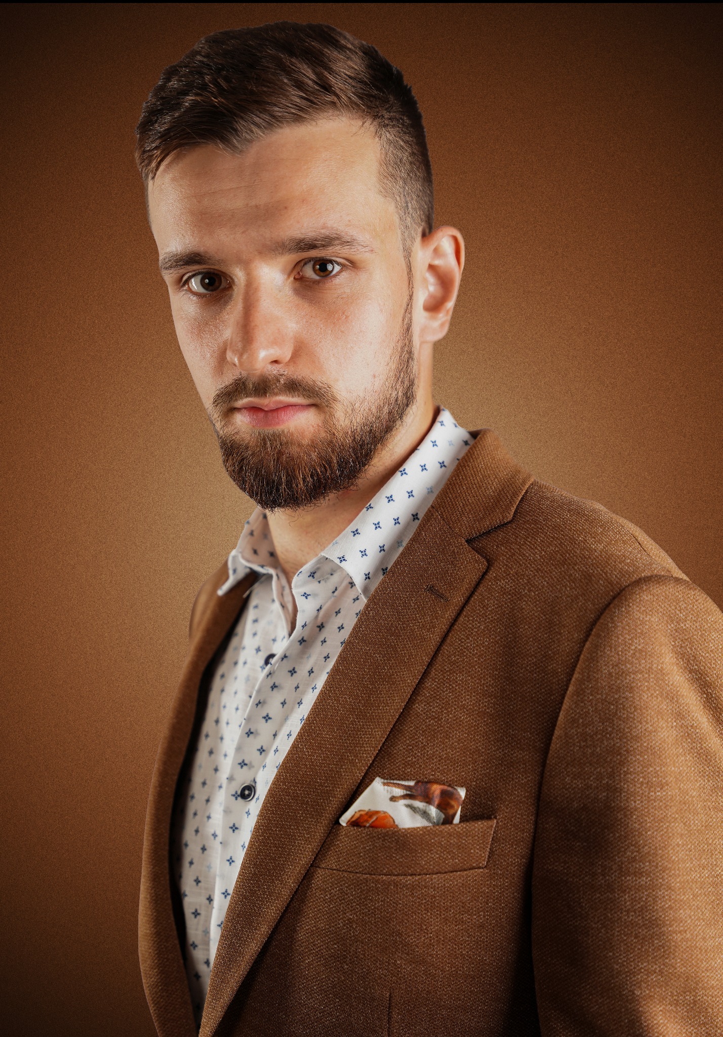 Portret młodego mężczyzny z brodą, w brązowym tweedowym garniturze i koszuli z wzorem, z poszetką w kieszonce, na jednolitym brązowym tle.