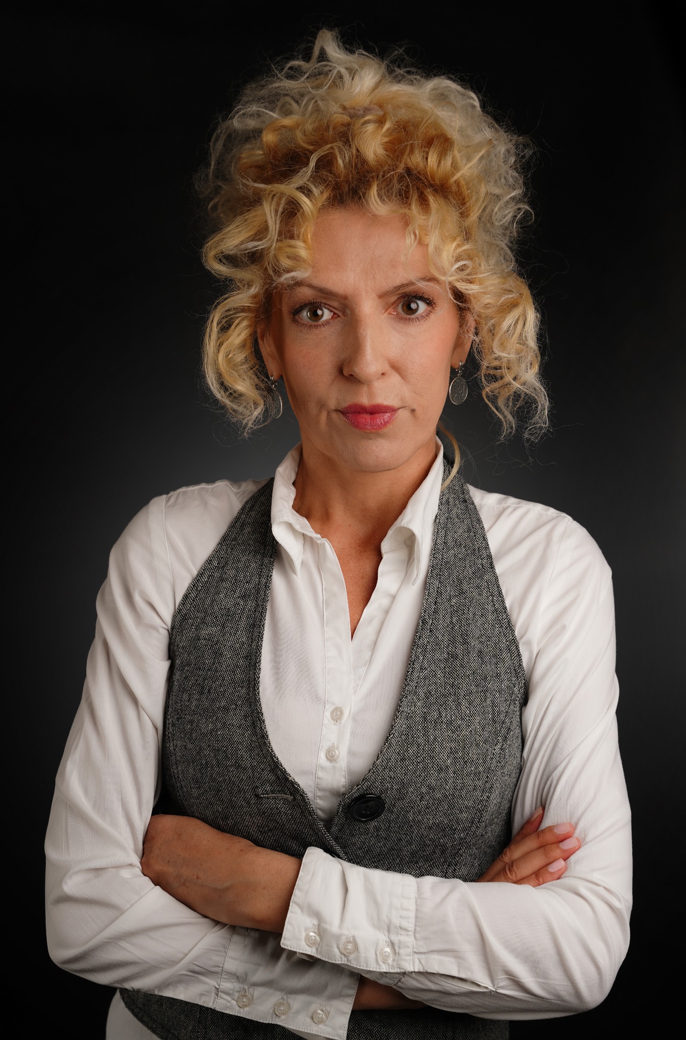 Portret kobiety z blond kręconymi włosami, ubrana w białą koszulę i szarą kamizelkę, z założonymi rękami na tle ciemnoszarego tła.