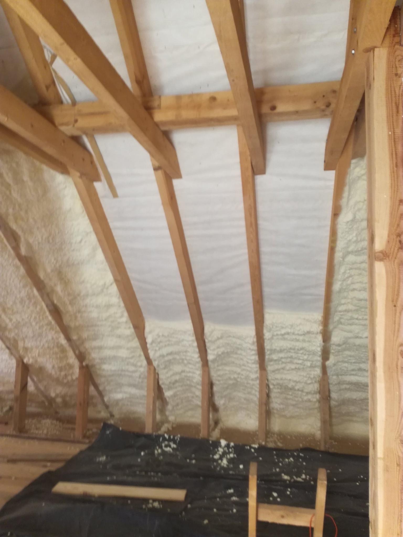 Poddasze w trakcie ocieplania pianką PUR, widoczne drewniane krokwie i biała pianka pokrywająca izolację, na podłodze czarna folia zabezpieczająca.