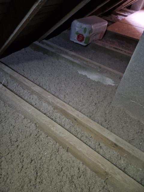 Poddasze z widoczną wełną mineralną między drewnianymi belkami stropowymi; na podłodze leży zapakowana paczka wełny.