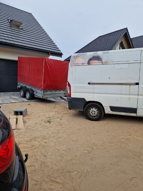 Biały van z przyczepą z czerwonym plandeką stoi na piaszczystym podwórku przed domem z ciemną dachówką i garażem.