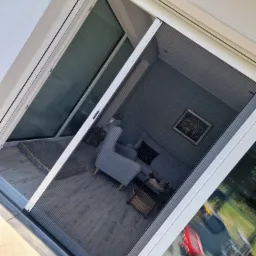 Zewnętrzna, plisowana siatka na owady w kolorze szarym, zamontowana w białej ramie okiennej, widok od wewnątrz, z odbiciem pokoju i ogrodu.