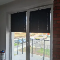 Dwa okna z czarnymi plisami, widok na balkon i okolicę, obok ściana z czerwonej cegły i wiaderka z farbą.