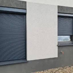 Dwa okna z opuszczonymi, grafitowymi roletami zewnętrznymi, elewacja budynku w odcieniach szarości i bieli, widoczny kran na ścianie.