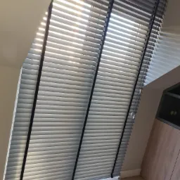 Szare żaluzje poziome z czarnymi taśmami, zamontowane w oknie dachowym, wpadające światło słoneczne tworzy cienie na ścianie.