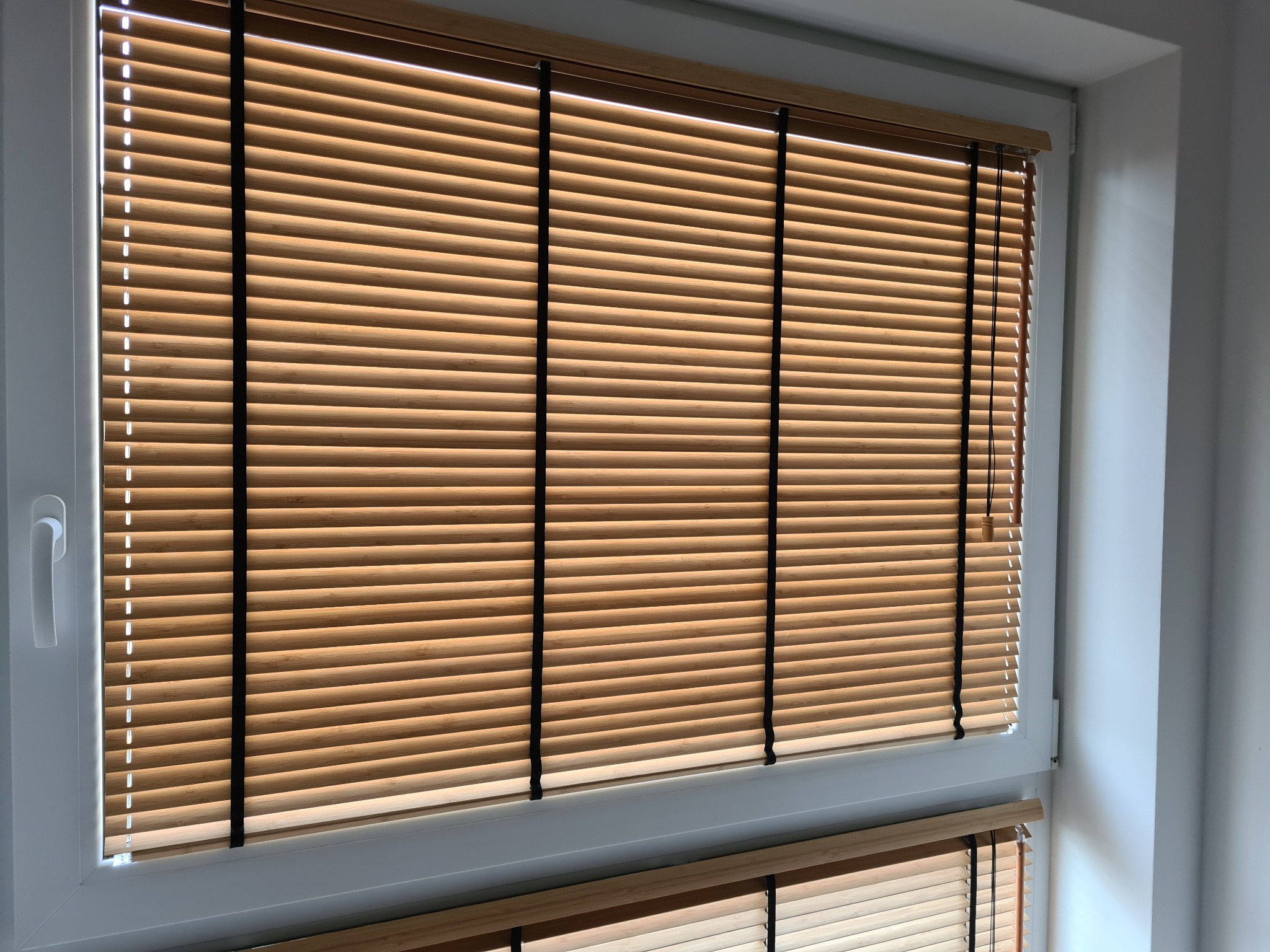 Jasne drewniane żaluzje poziome w białej ramie okiennej, częściowo zasłonięte, z widocznymi sznurkami po prawej stronie i fragmentem drugiej żaluzji u dołu.