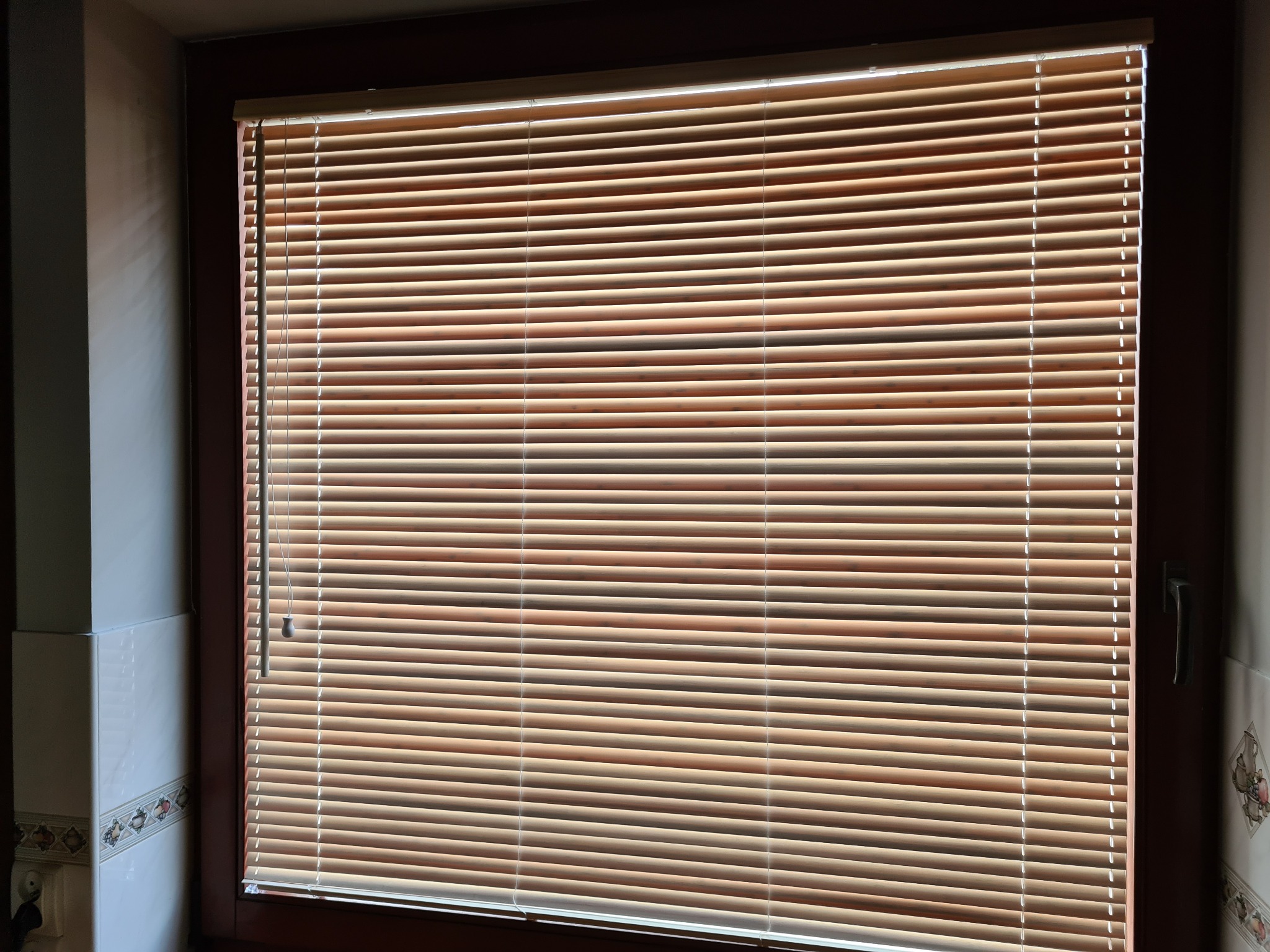 Zamknięte poziome żaluzje w odcieniach brązu w oknie z ciemną ramą, widoczny fragment ściany z dekoracyjnymi płytkami.