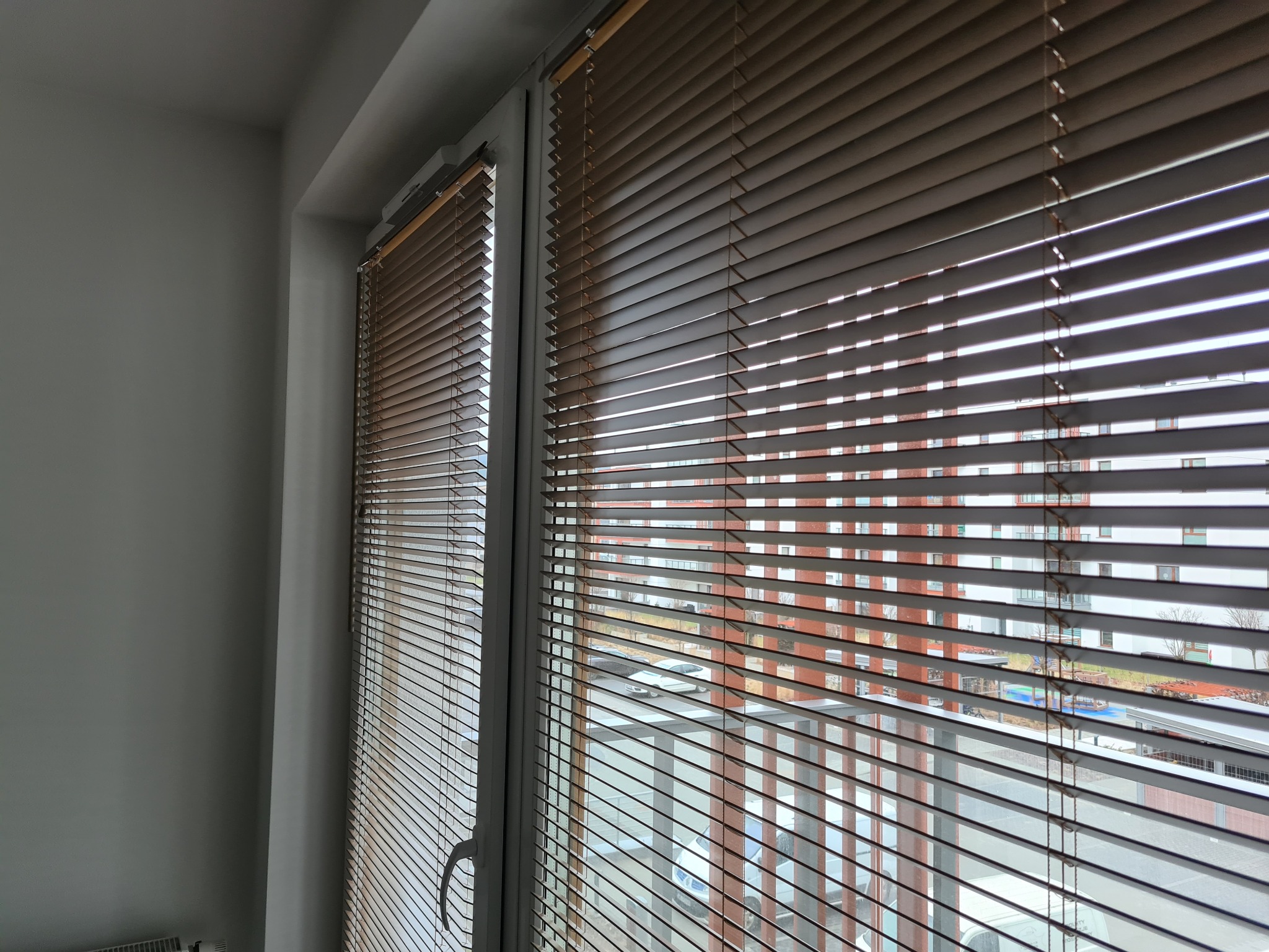 Jasne, poziome listwy w oknie, częściowo zasłonięte, widok na budynek mieszkalny z placem zabaw.