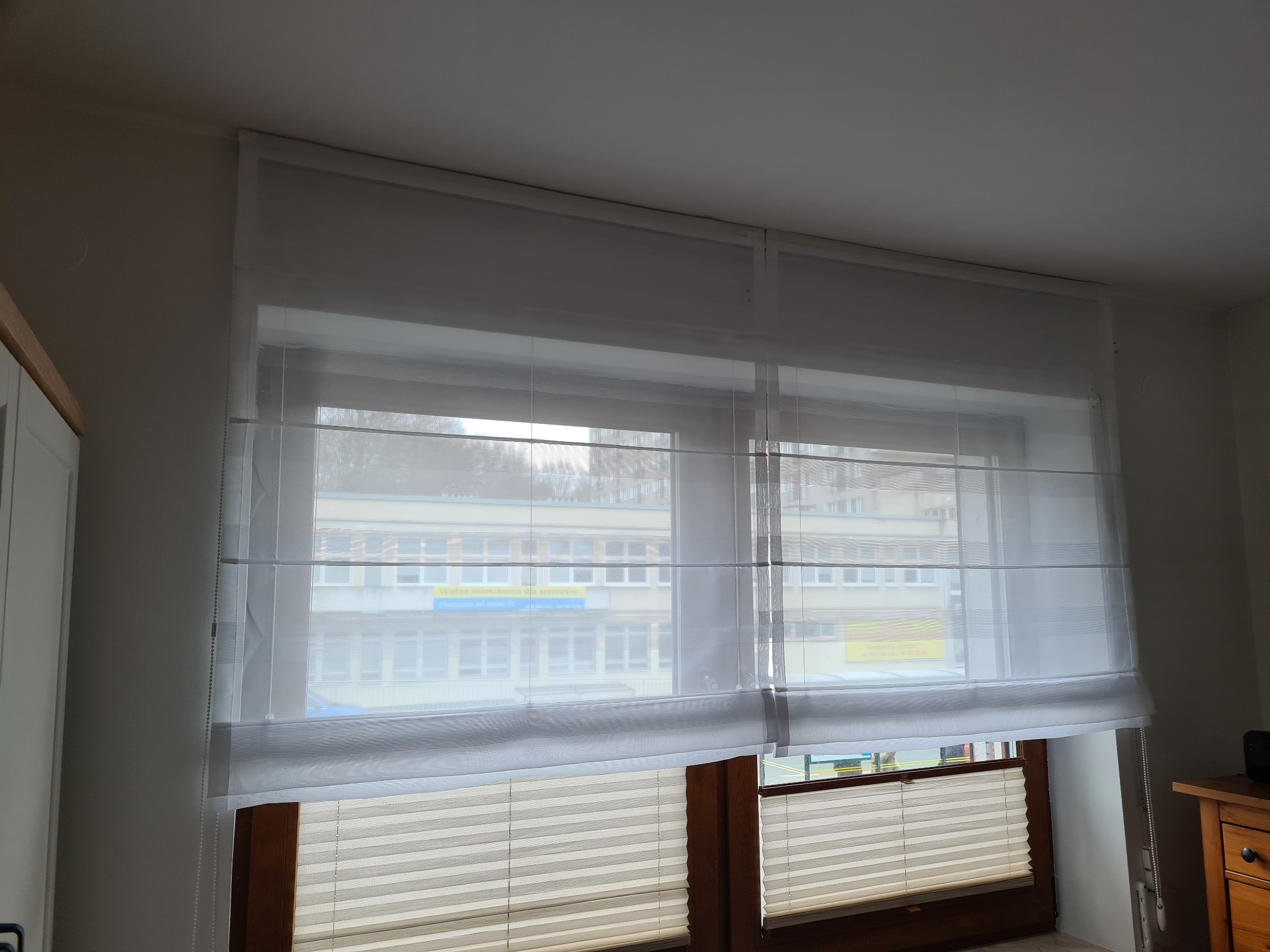 Dwa okna z zamontowanymi roletami rzymskimi z jasnej, prześwitującej tkaniny oraz plisami w dolnej części okien. Na zewnątrz widoczny budynek.