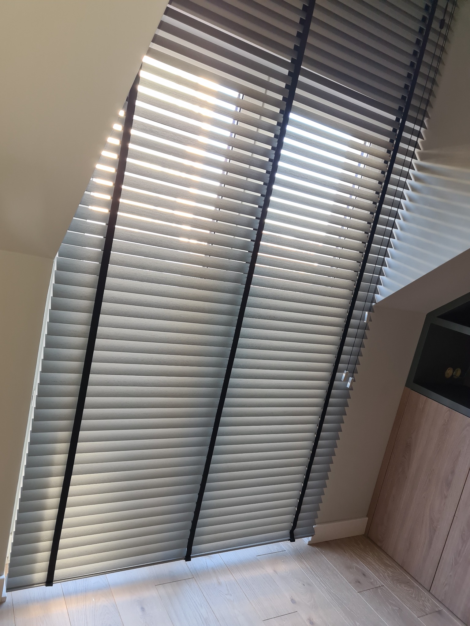 Szare żaluzje poziome z czarnymi taśmami, zamontowane w oknie dachowym, wpadające światło słoneczne tworzy cienie na ścianie.