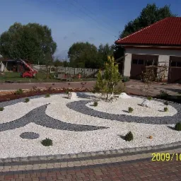 Ogród z dekoracyjnym wzorem z białych i szarych kamieni, niewielkimi krzewami i młodymi drzewkami, widok z poziomu gruntu na dom z czerwonym dachem w tle.