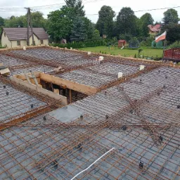 Zbrojenie płyty fundamentowej na etapie przygotowania do zalewania betonem, widoczne drewniane szalunki i dystanse, w tle domy jednorodzinne i zieleń.