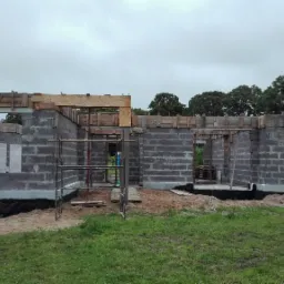Wznoszenie ścian z bloczków betonowych na fundamentach, z drewnianym szalunkiem nad otworami okiennymi i drzwiowymi, widok na plac budowy i zieleń w tle.