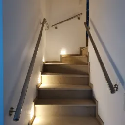 Schody z kamienia z zamontowanymi, stalowymi poręczami oświetlone punktowymi lampkami LED w ścianie.