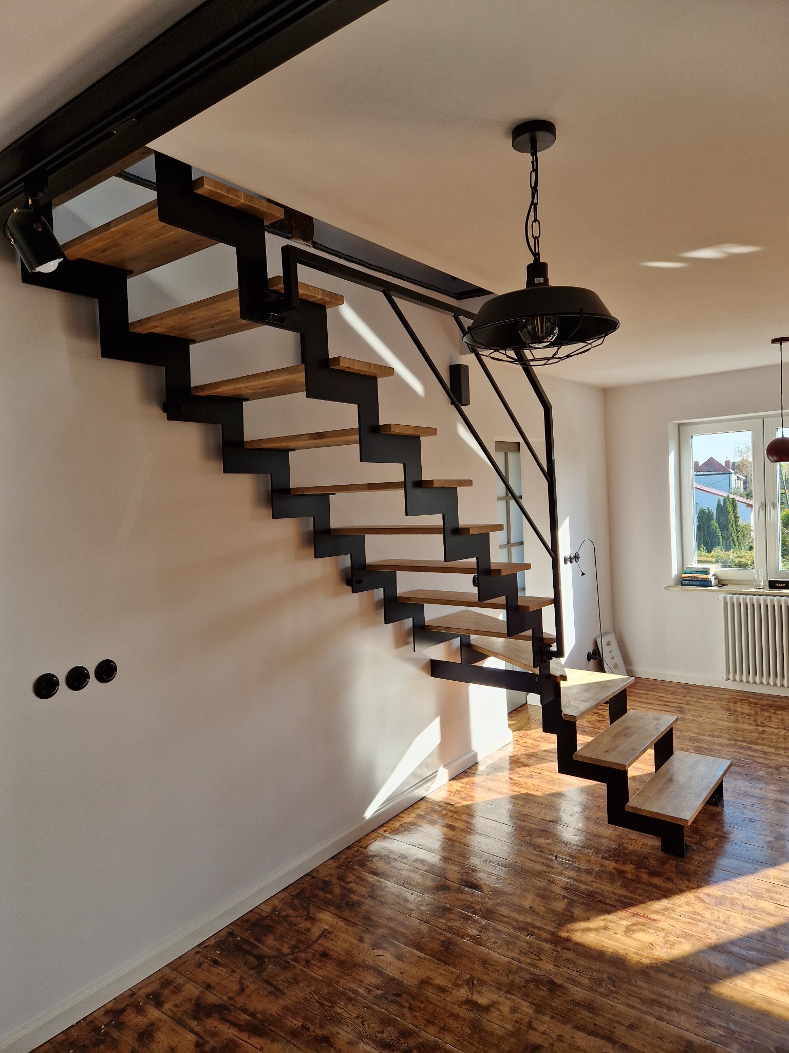 Nowoczesne schody z drewnianymi stopniami i czarną metalową konstrukcją, oświetlone padającym przez okno światłem, na tle drewnianej podłogi.