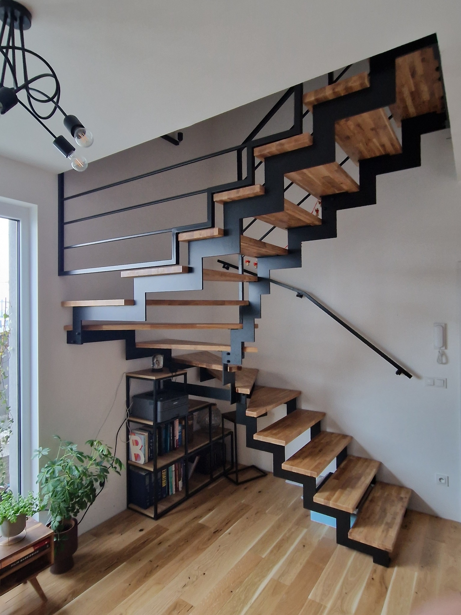 Nowoczesne schody wspornikowe z drewnianymi stopniami i czarną metalową konstrukcją, półka na książki pod schodami, widok z dołu.