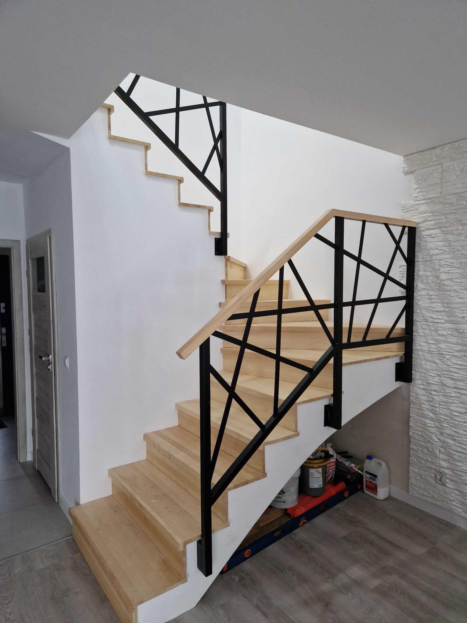 Drewniane schody z czarną, geometryczną balustradą metalową, widok z dołu, pod schodami widoczne narzędzia i materiały budowlane.