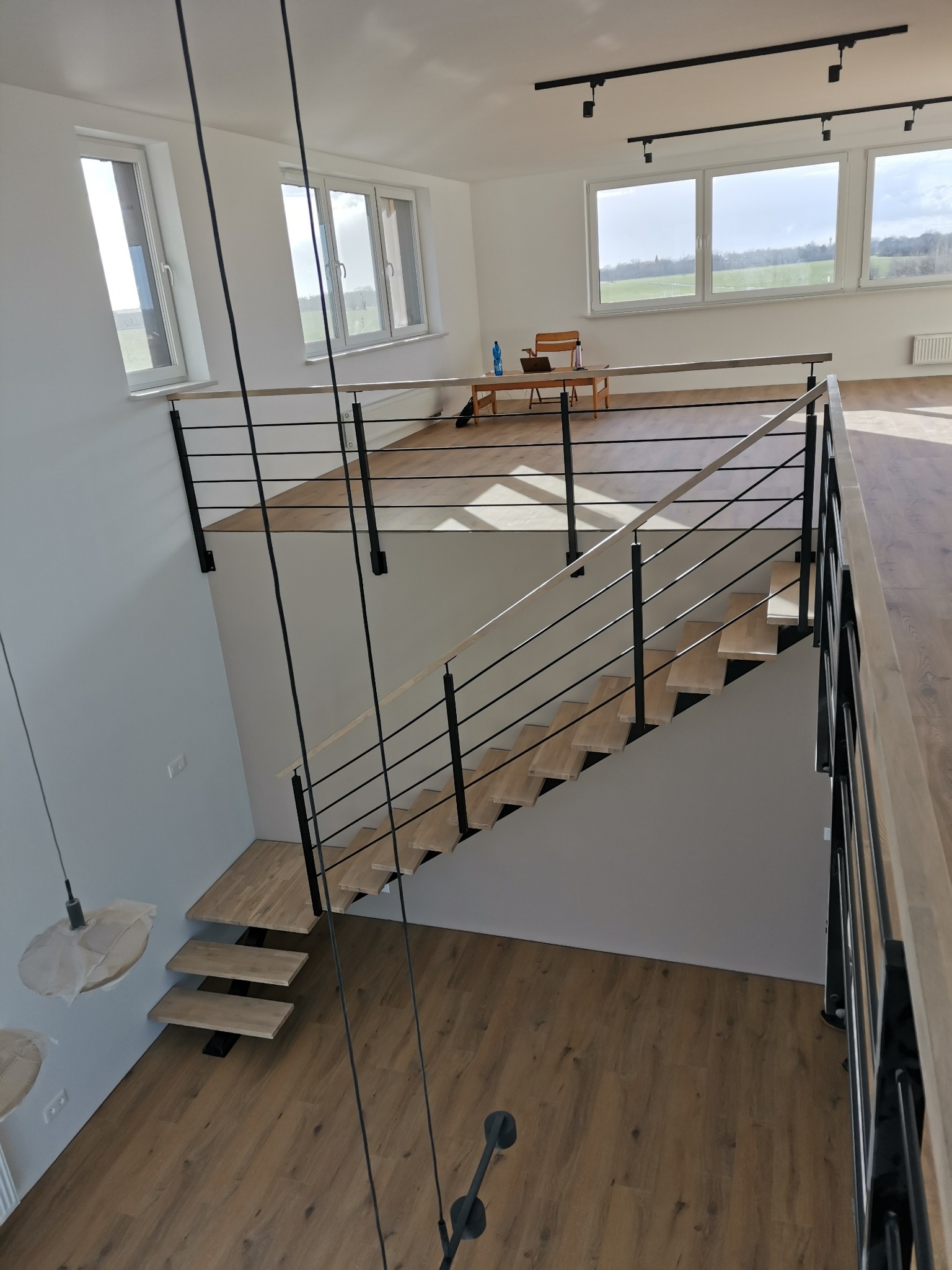 Wnętrze domu z drewnianymi schodami, czarnymi metalowymi elementami konstrukcyjnymi i poziomą balustradą z jasnego drewna, widok z góry na schody i fragment pomieszczenia z oknami.