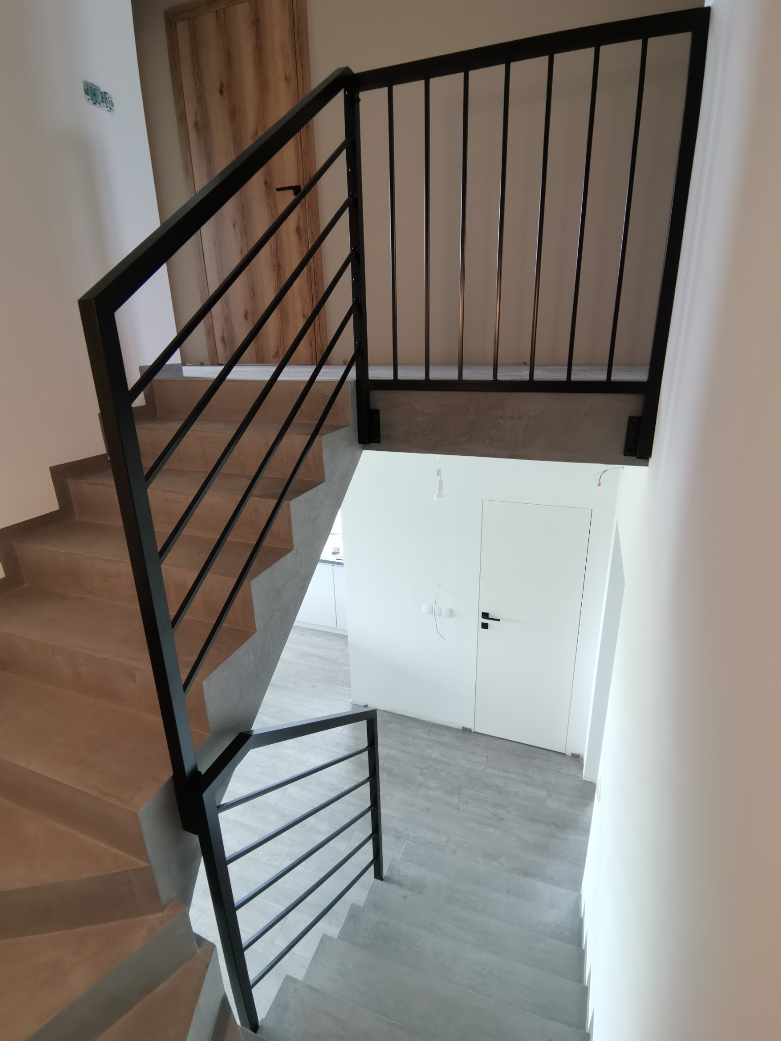 Wnętrze z betonowymi schodami i czarną, minimalistyczną balustradą z poziomych prętów, widok z góry na dół, z widocznymi elementami wykończenia.