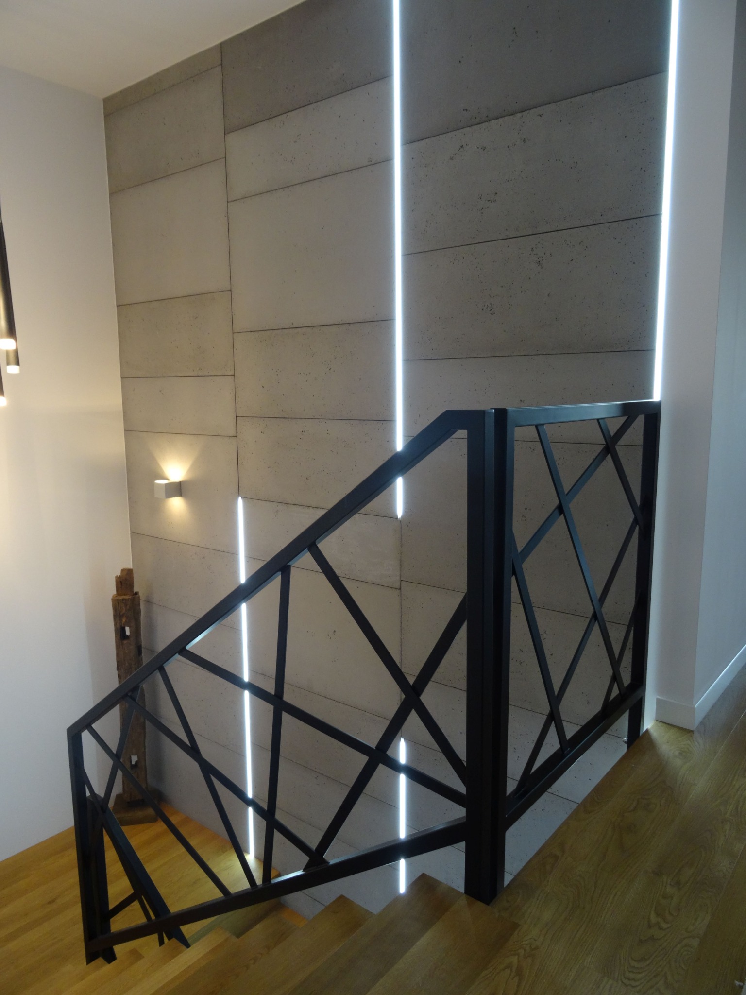 Czarna, geometryczna konstrukcja zabezpieczająca schody z drewnianymi stopniami, na tle betonowej ściany z pionowymi paskami oświetlenia LED.