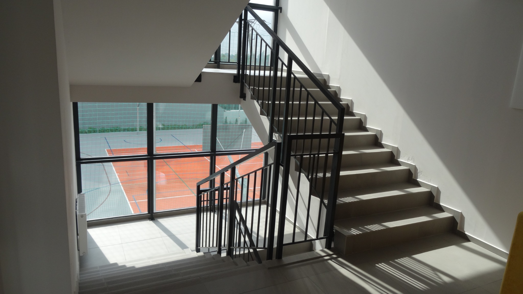 Klatka schodowa z czarną, metalową konstrukcją zabezpieczającą, widok z góry z oknem na boisko sportowe w tle.