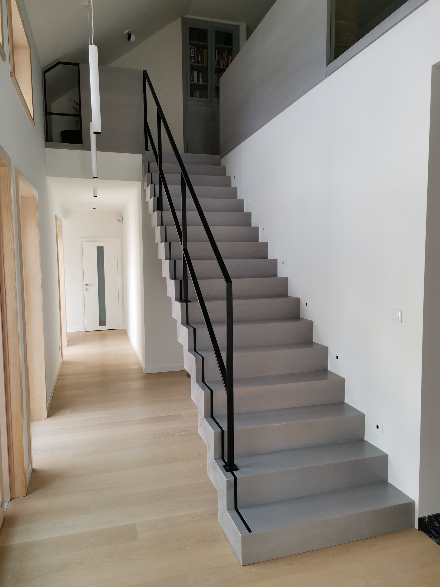 Nowoczesne, betonowe schody z czarną, metalową balustradą w jasnym wnętrzu z drewnianą podłogą i minimalistycznym oświetleniem.