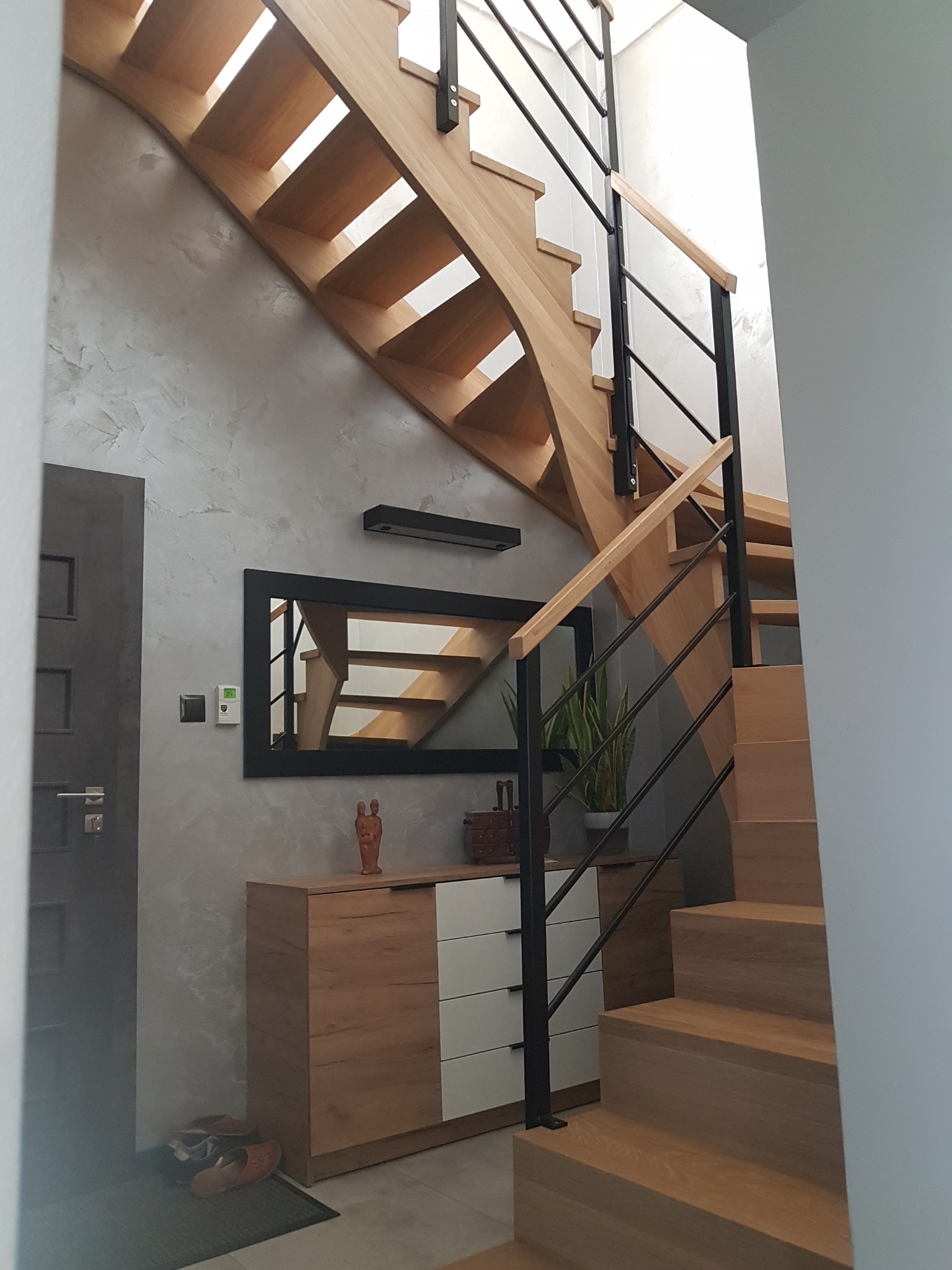 Drewniane schody z czarną, minimalistyczną balustradą, widoczne z dołu, z szafką i dekoracjami w tle, odbijające się w lustrze.