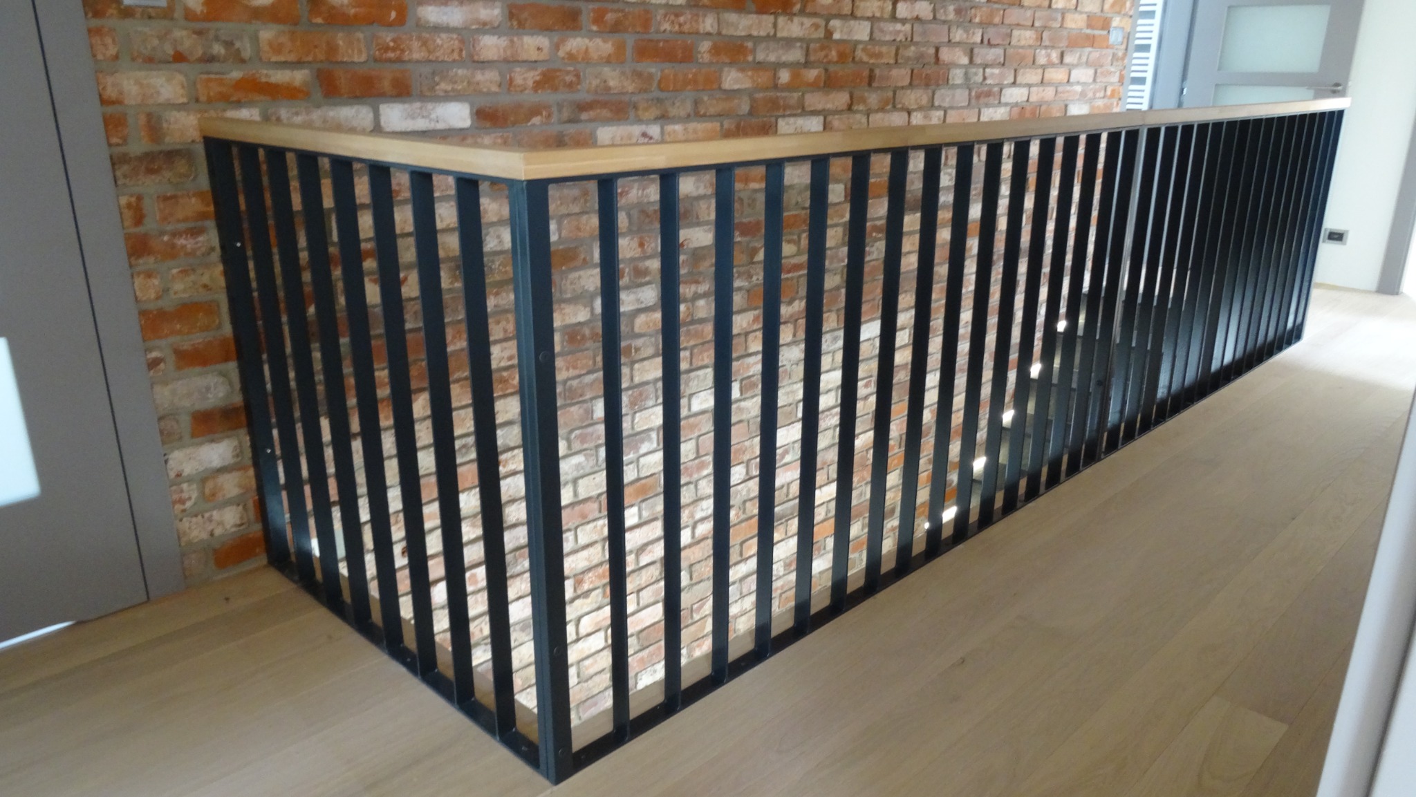 Nowoczesna balustrada z czarnych pionowych prętów i drewnianym blatem, zamontowana przy schodach w pomieszczeniu z ceglaną ścianą